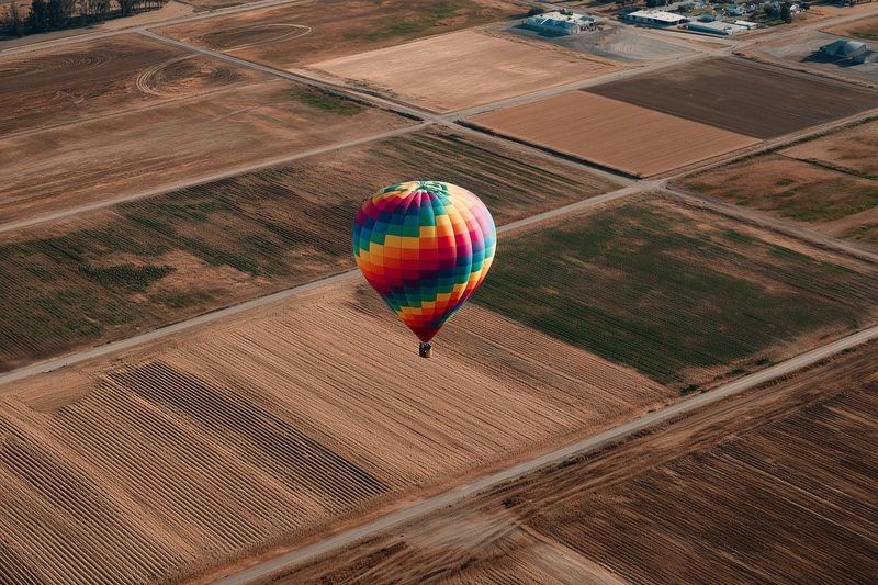 Colorful hot air balloon floating above a patchwork of agricultural fields at sunrise, casting a soft shadow over dirt roads and crops in a serene aerial landscape scene.