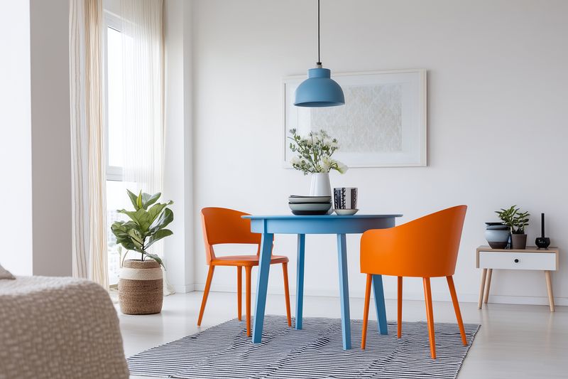 Bright modern dining area with a blue round table and two orange chairs on a patterned rug, lit by a hanging pendant lamp and soft natural light from a large window.