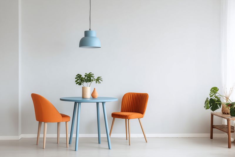 Minimal modern dining nook with a round blue table, two orange upholstered chairs, hanging pendant lamp and potted plants against a clean white wall, bright interior.