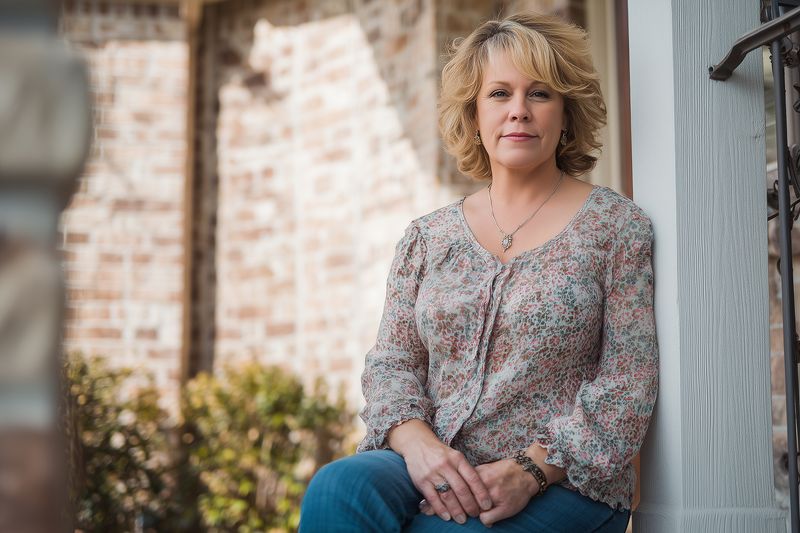 Portrait of a middle aged woman sitting on a porch step in casual clothing, relaxed pose and soft natural light with shallow depth of field conveying quiet confidence.