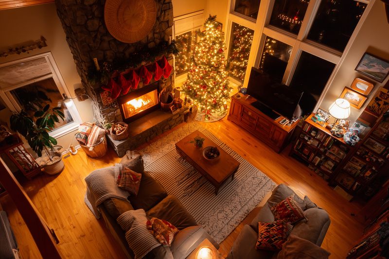 Cozy living room decorated for Christmas with glowing tree lights, roaring fireplace, stockings on the mantel, warm wooden floors and festive pillows creating holiday charm.