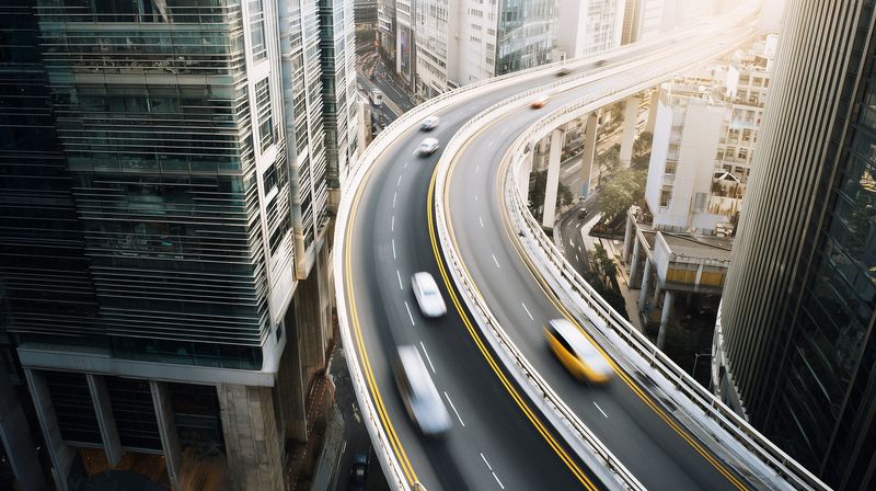 Aerial perspective of a sweeping elevated highway curving between highrise buildings in a bustling modern city, with streaking vehicles and motion blur conveying speed and urban energy.