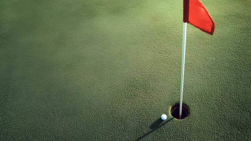 Close-up of a golf hole with a red flagstick and a white golf ball resting on the green turf, sunlight casting a sharp shadow across the textured putting surface.