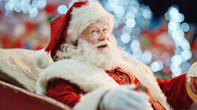 An elderly man dressed in a traditional red holiday costume with white fur trim and fluffy beard smiling warmly while seated on a sleigh amid soft festive bokeh lights in background.