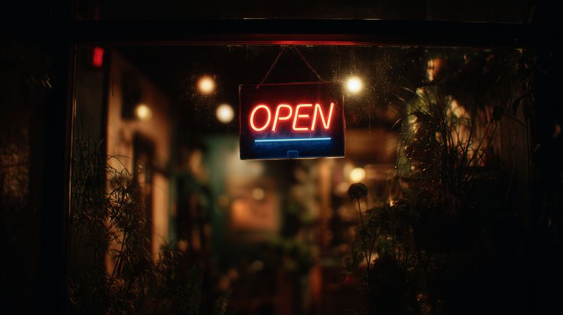 Warm glowing neon OPEN sign hanging in a storefront window at night, blurred interior lights and plants create a cozy, inviting atmosphere for passing customers and urban evening scenes.