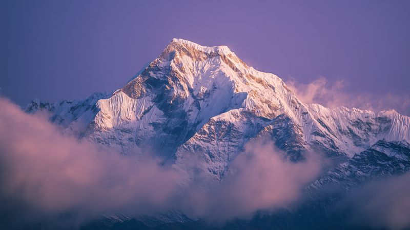 Snow capped mountain peak bathed in soft purple dawn light, dramatic clouds swirling around the summit creating a serene and majestic alpine landscape full of texture and contrast.