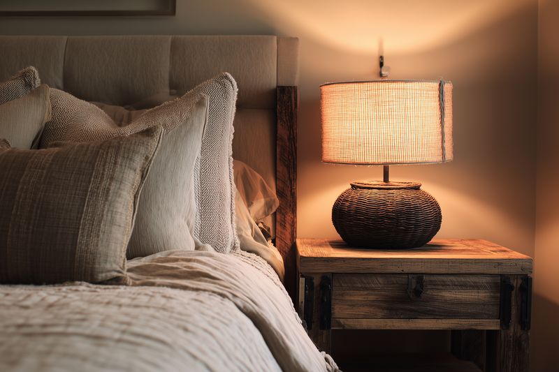 Warm bedside lamp illuminates a cozy neutral bedroom setting with textured pillows, layered bedding and a rustic wooden nightstand creating inviting, serene sleeping space atmosphere.