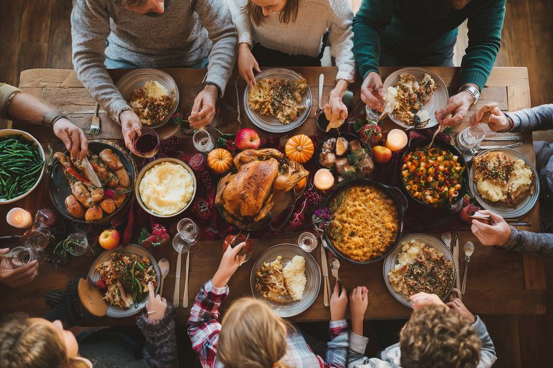 Family gathered around a wooden table sharing a warm festive meal with a golden roasted turkey, side dishes, candles and autumn decorations in a cozy communal holiday dinner scene.