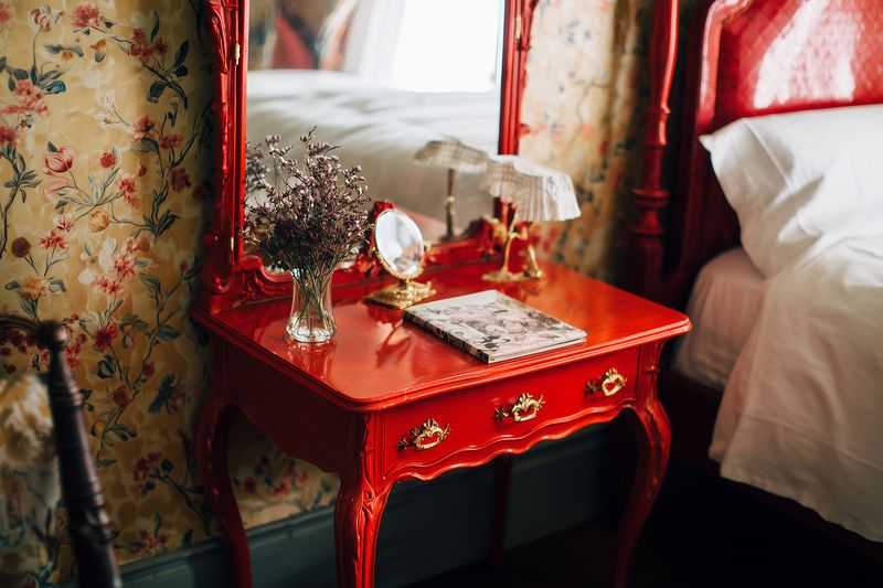 Vintage red vanity table with ornate mirror, decorative lamp, dried flowers in a glass vase and a patterned notebook beside a cozy bed, set against floral wallpaper.