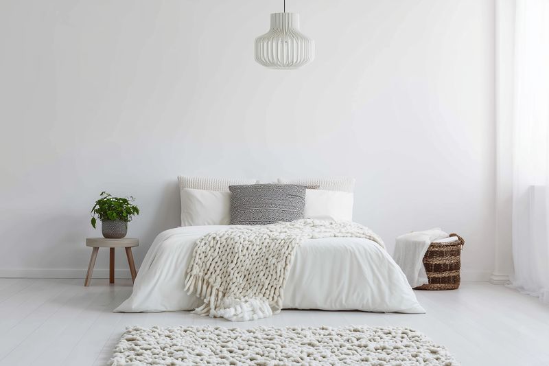 Minimalist bright bedroom interior with a neatly made white bed, textured throw and rug, soft pillows, woven basket and potted plant, illuminated by natural light and pendant lamp.