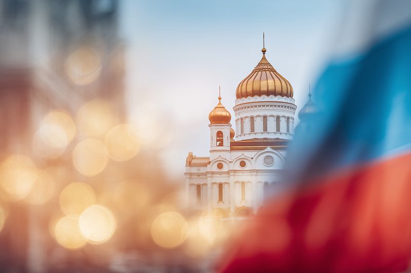 Sunlit golden domes of an ornate cathedral rise behind a softly blurred foreground, with warm bokeh highlights and a waving tricolor flag suggesting cultural heritage, ceremony and timeless reverence.