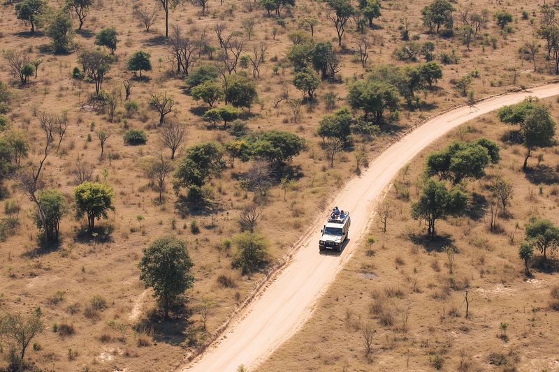 Aerial view of a safari vehicle driving on a winding dirt road through dry savanna with scattered trees, conveying remote landscape, adventure and wildlife observation mood.