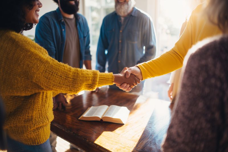 Diverse adults in a casual meeting share a friendly handshake over a wooden table with an open book, warm sunlight and smiling faces conveying trust and collaborative spirit.