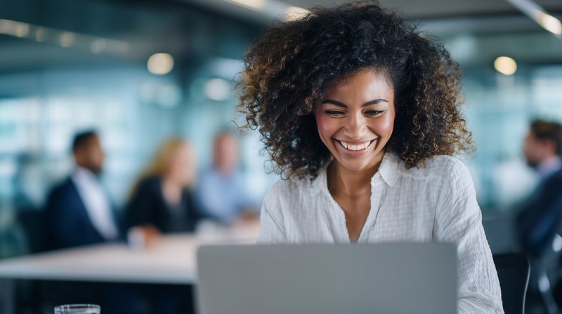 Smiling young professional woman working on a laptop in a modern office environment, focused and engaged with natural light and blurred colleagues in the background.