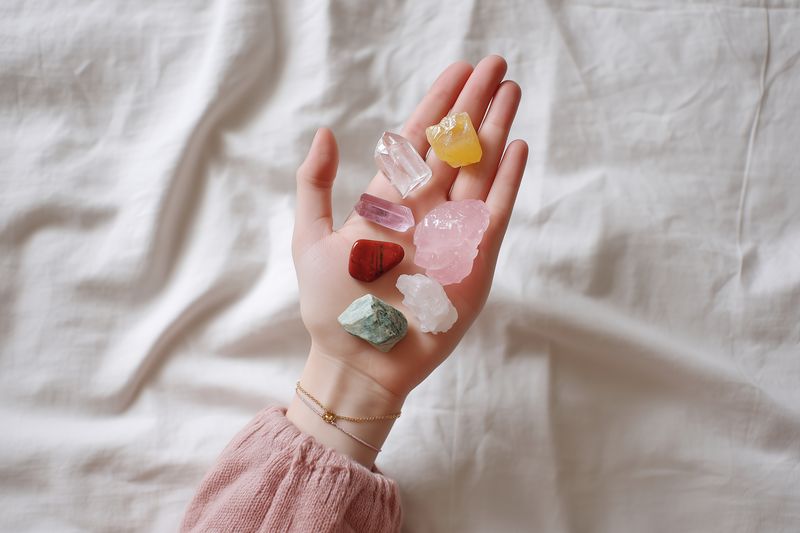 Close-up of a hand holding assorted raw healing crystals and stones on a soft fabric background, highlighting pastel colors, natural textures, and a calm, minimalist boho aesthetic.