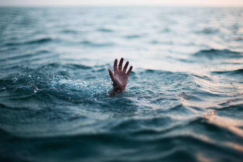 A solitary hand emerges from turbulent ocean water, fingers outstretched above splashing waves, creating a dramatic scene that conveys struggle, urgency, and raw emotional tension.