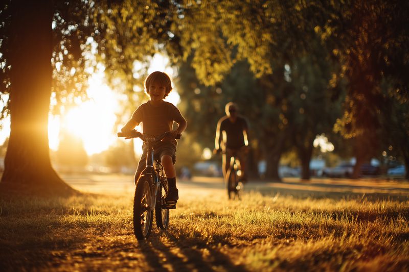 Young child riding a bicycle along a sunlit park pathway at golden hour, with an adult following among trees and warm rays creating long shadows and peaceful summer atmosphere.
