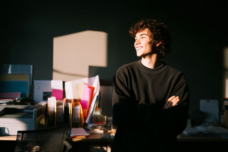 young man smile in office workspace by desk with sunlight on face, curly hair and casual sweater, creative in studio with documents folders and modern work environment