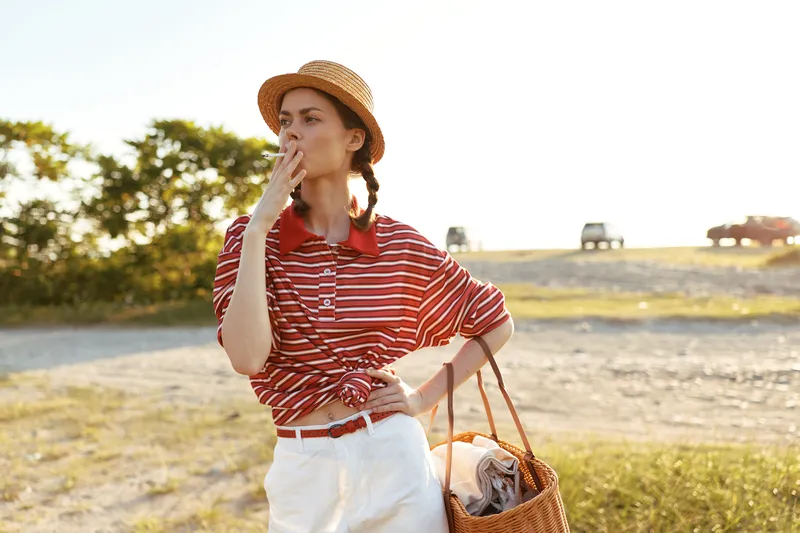 Young woman, lifestyle, straw, hat, cigarette, standing, outdoors. She wears striped shirt and white pants while posing in nature. Sunset lighting concept for leisure portrait.