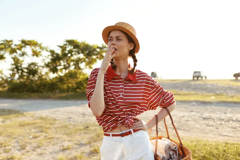 Young woman enjoying lifestyle outdoor smoking a cigarette in a sun hat. Female model with braids in summer clothes standing on a field during sunset. Concept of leisure and summer vacation.