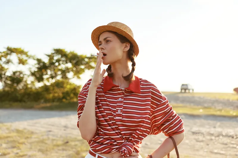 Young woman in a straw hat and striped shirt yawns while standing outdoors. Authentic lifestyle portrait of a girl posing with braided hair on a sunny summer day.