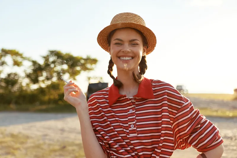 Young woman in a straw hat and striped shirt smiles while holding a cigarette during a summer outdoor lifestyle session. The golden sunset light illuminates her happy face and casual clothing.