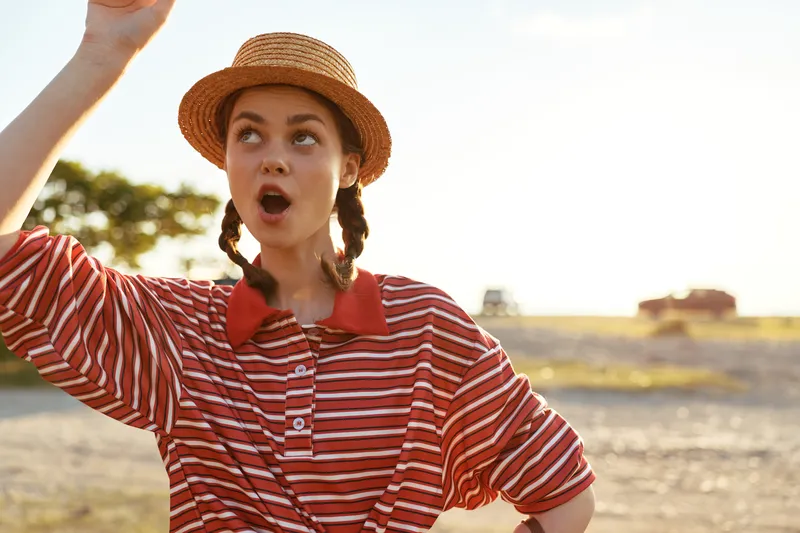 Young woman wearing a straw hat and a red striped shirt looking up with a surprised expression. Lifestyle portrait in the summer sun outdoors. Concept for summer vacation and travel joy mood.
