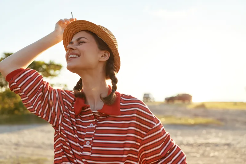 Smiling woman wearing a straw hat and striped shirt enjoying sunny outdoor lifestyle. Golden hour light creates a happy mood. Concept of freedom, summer, happiness, and travel relaxation.