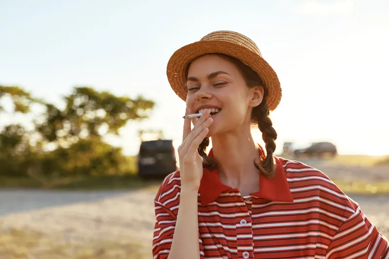 Young woman with braided hair wearing a straw hat smokes a cigarette outdoors. The authentic lifestyle portrait captures a carefree expression during a sunny day in a relaxed atmosphere.