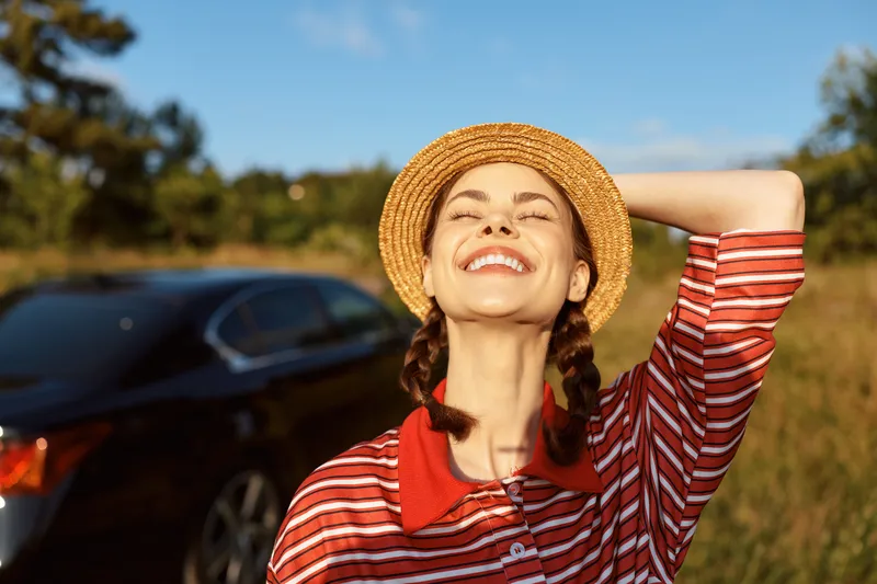 Happy woman lifestyle portrait in straw hat standing near car outdoors. She enjoys summer nature and sunlight, eyes closed with smile. Concept of travel and freedom.
