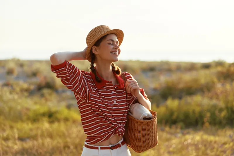 A smiling young woman in a straw hat and striped shirt carrying a wicker basket in a field at sunset. A happy lifestyle concept capturing outdoor summer leisure and relaxation.