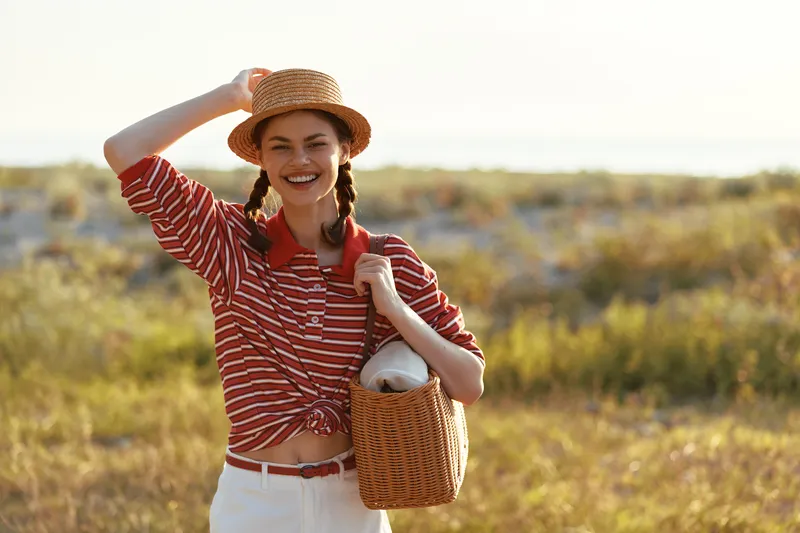 Young woman in a straw hat and striped sweater laughs during a summer walk in nature. She carries a wicker bag while enjoying a warm sunny day in this authentic portrait.
