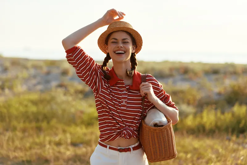 Happy woman enjoys lifestyle during summer vacation at the beach. She wears straw hat and striped shirt with bag. Outdoor portrait capturing joy, leisure, travel, sunny day.