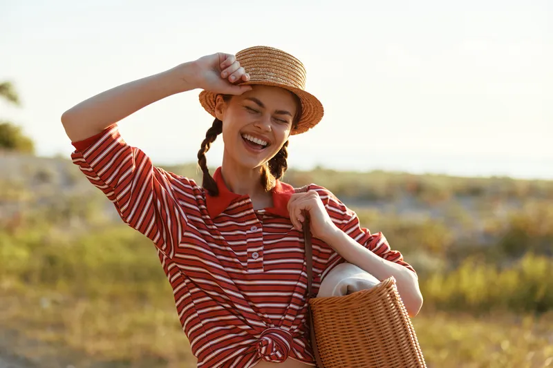 Happy young woman with pigtails wearing straw hat and striped shirt carrying wicker bag walks on sunny nature background enjoying leisure time and lifestyle during summer vacation.