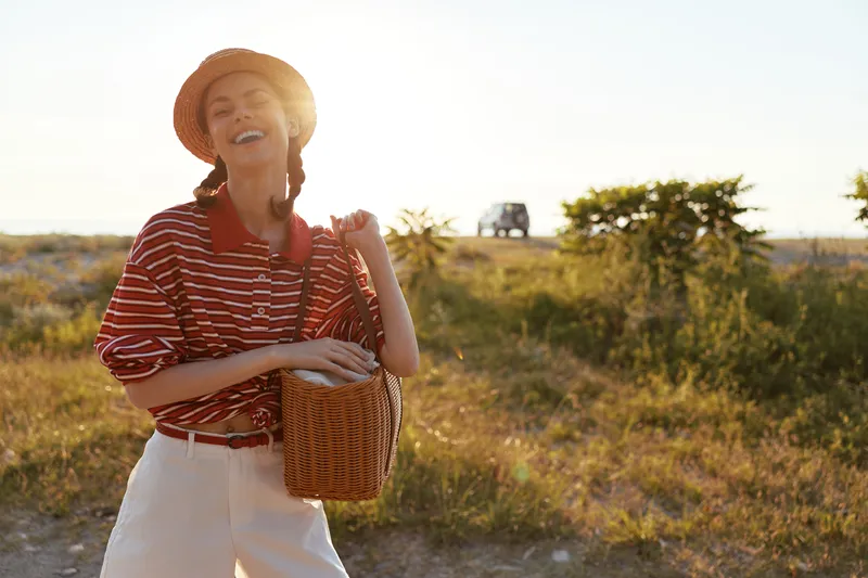A happy woman lifestyle portrait in a straw hat holding a wicker bag outdoors. Sunny summer meadow background, enjoying vacation, leisure, and travel in casual attire.