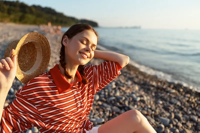 Young woman with braids holding straw hat enjoying sunny day on pebble beach by sea, concept of summer vacation and joyful lifestyle for authentic travel content.