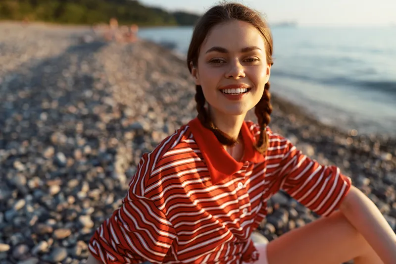 Smiling woman lifestyle beach portrait outdoors. Casual clothing, sunset natural lighting, sunny sea pebbles background. Happy vacation, leisure, and summer vibe concept.