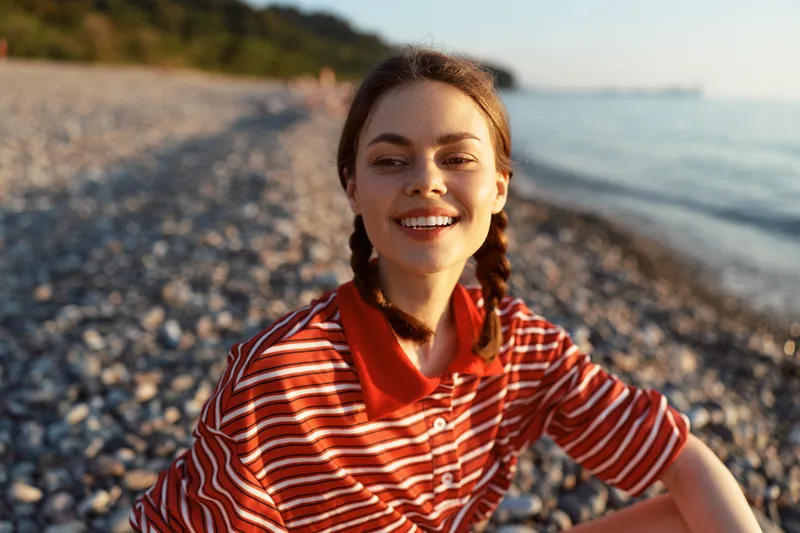 A smiling young woman lifestyle portrait on a pebble beach at sunset. She wears a striped shirt with braids while enjoying the summer sea breeze. Concept for travel.