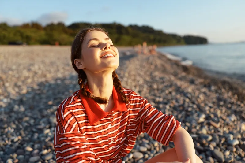 Young woman enjoying lifestyle on a pebble beach at sunset. She is smiling with eyes closed feeling the sun beams. Concept for leisure, relaxation, and serenity.