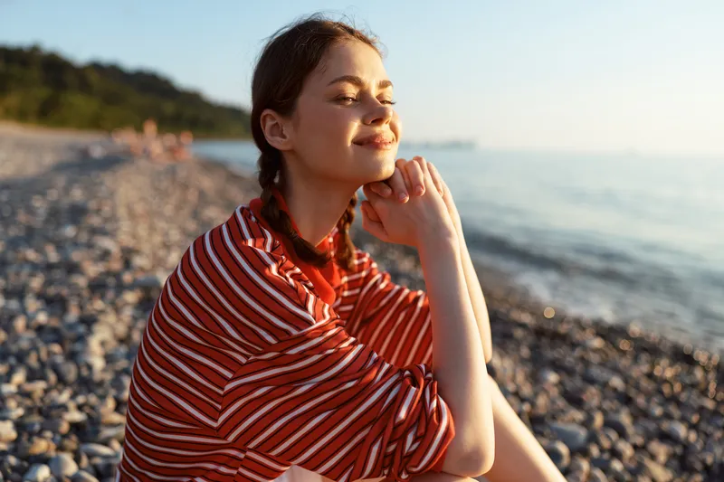 Young woman enjoying lifestyle during a relaxing summer sea sunset. Portrait of a happy lady sitting on a rocky beach by the ocean. Warm sunlight, calm mood, natural look.