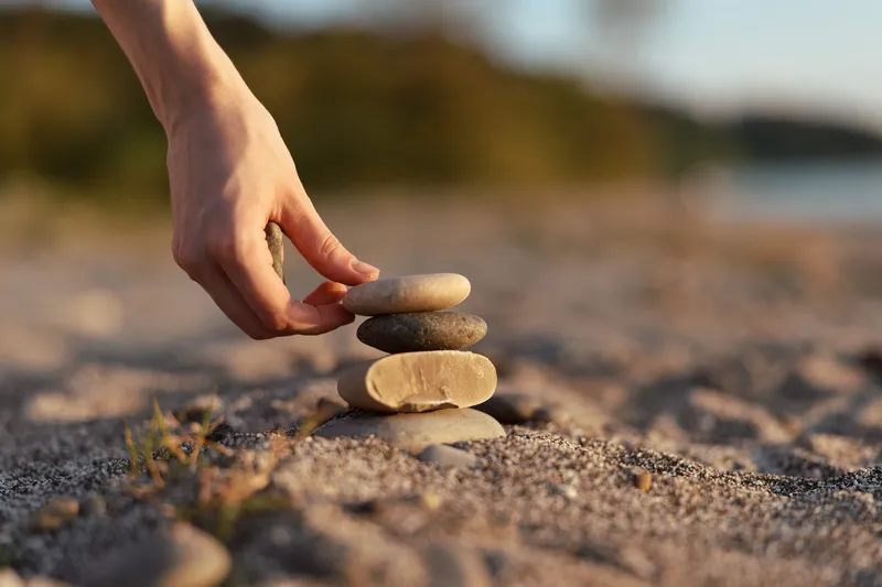 A woman builds a stack of stones on a beach at sunset, demonstrating lifestyle balance, meditation, and zen. Concept for mindfulness, peace, and meditation in nature outdoor.