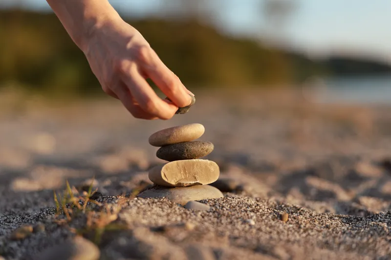 A woman builds a zen stone balance tower on a sandy beach during sunset. This peaceful scene captures the essence of mindfulness, relaxation and lifestyle balance.