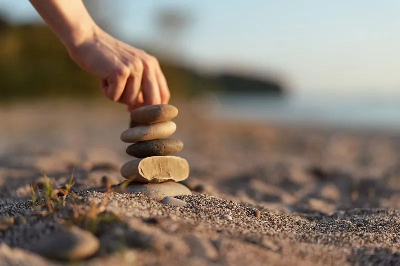 Hand stacking smooth stones into a balance tower on a sandy beach during sunset, representing mindfulness, peace, and internal harmony in a serene coastal lifestyle environment.