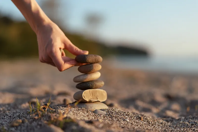Woman lifestyle concept showing a hand stacking natural stones on a sandy beach at sunset. The balancing stones represent harmony, peace, zen, and meditation practice.