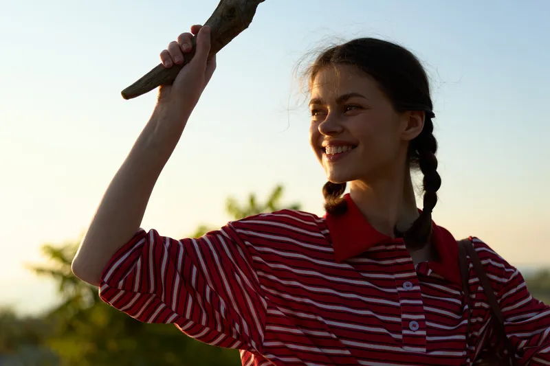 Smiling woman posing outdoors for a lifestyle portrait. She holds a wooden stick at sunset. Summer freedom, joy, and natural beauty in a bright and happy candid moment.