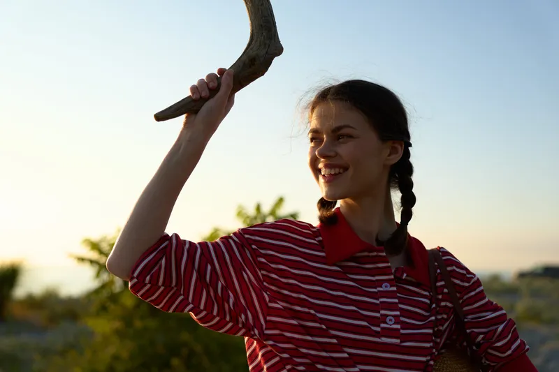 Smiling woman holding a wooden stick outdoors. Lifestyle portrait captures natural joy and sunset light. Concept of outdoor travel, happiness, vacation, summer, freedom, nature.