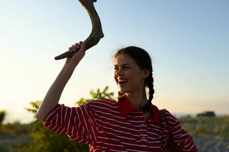 Young woman enjoying lifestyle on the beach holding a piece of driftwood. The happy girl smiles during sunset outdoor. Authentic portrait capturing a joyful summer moment in nature near ocean.