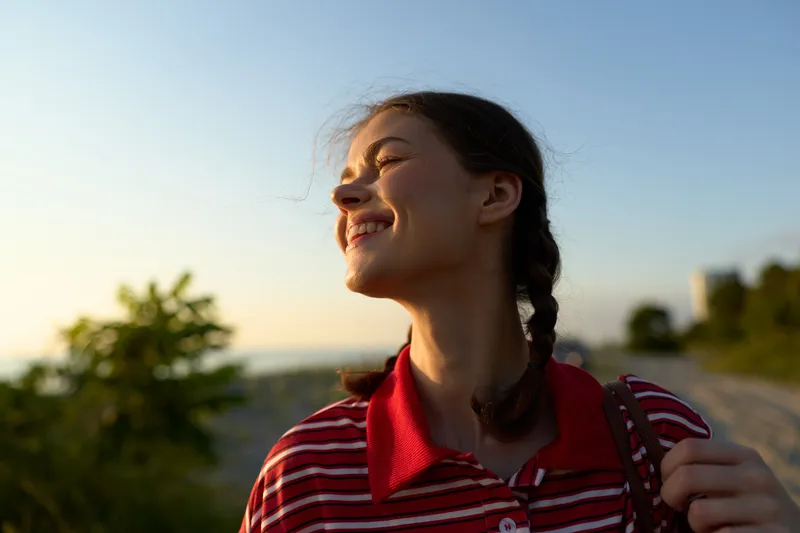 Smiling young woman with braided hair enjoys warm sunlight outdoors at sunset while wearing a striped polo shirt. Authentic portrait captures a happy, peaceful lifestyle moment in nature scenery.