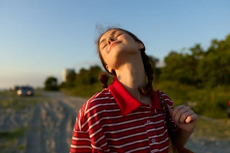 A happy young woman enjoying sunset lifestyle, feeling freedom, peace and joy outdoors. She closes her eyes, smiling under the warm golden summer light in nature.