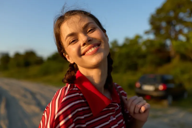 Smiling young woman with braided hair enjoys a sunny summer road trip outdoors. The cheerful girl in a striped shirt poses for a portrait captured in a warm natural daylight.
